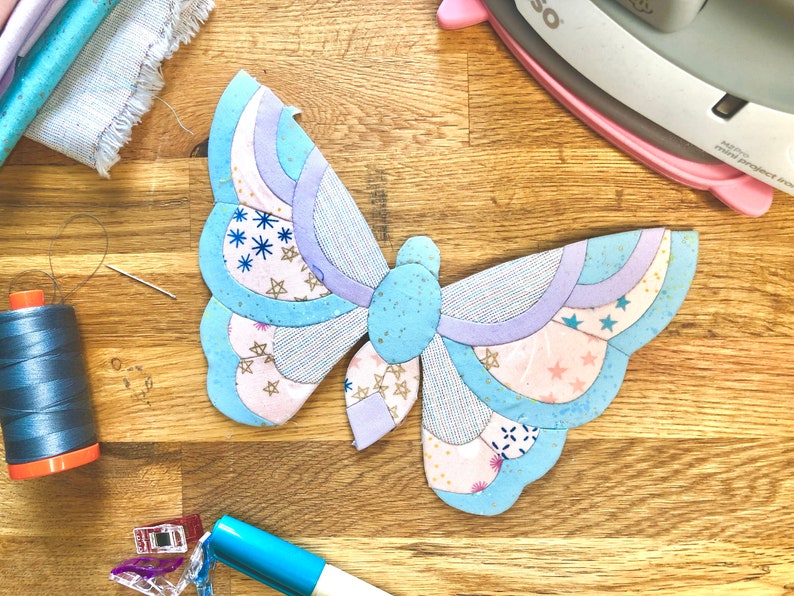 May include: A blue and purple butterfly-shaped fabric applique with a pink and white patterned design. The applique is on a wooden surface with sewing supplies.