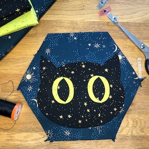 May include: A black cat with yellow eyes is appliqued onto a blue fabric hexagon with gold stars. The fabric is surrounded by other fabric scraps and sewing tools.