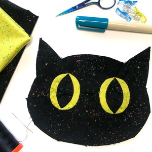 May include: Black fabric cat face with yellow eyes, a needle and thread, a spool of black thread, a blue marker, and a pair of silver scissors on a white surface.