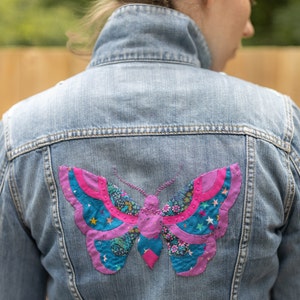May include: A denim jacket with a colorful butterfly patch sewn on the back. The butterfly is made of fabric scraps in shades of pink, blue, and green.
