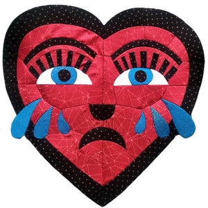 May include: A heart-shaped fabric art piece with a red face, blue eyes, and black eyelashes. The heart has blue teardrops and a black border with gold stars. The face has a sad expression.