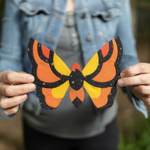 May include: A person is holding a large, colorful butterfly made of fabric. The butterfly has orange, yellow, and black wings with a pattern of stars.