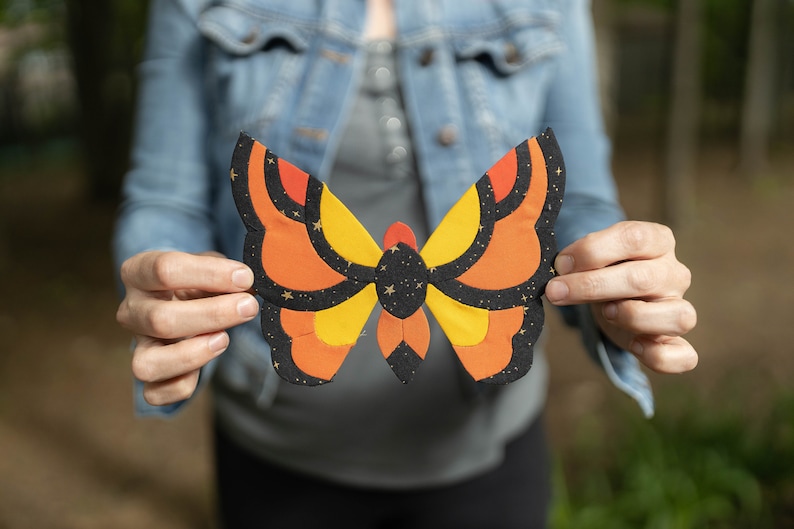May include: A person is holding a large, colorful butterfly made of fabric. The butterfly has orange, yellow, and black wings with a pattern of stars.