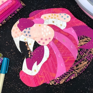 May include: A pink and gold fabric applique of a tiger's head with a black background. The tiger's mouth is open and its teeth are visible. The fabric is patterned with stars, moons, and geometric shapes.