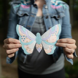 May include: A person holds a blue and pink butterfly-shaped paper craft in front of them. The butterfly is made of different fabric scraps and has a delicate, whimsical design.