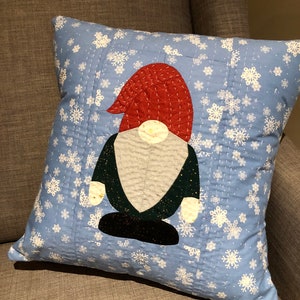May include: A blue pillow with a white snowflake pattern. A red, green and black gnome is appliqued on the pillow.