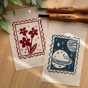 以下が含まれることがあります： 赤色の花の模様と青色の惑星と星が描かれた、テクスチャード加工された紙に印刷された2つのリノカットプリント。木製のハンドルが付いた2つのPower Grip彫刻ツールが見えます。