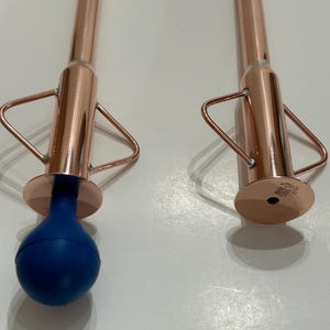 May include: Two metallic copper-colored tools with long cylindrical shafts and triangular side handles. One tool features a blue spherical bulb at the base, while the other has a flat circular base with small engraved text reading HURL.