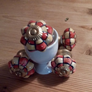 May include: Four ceramic cabinet knobs with a yellow, red and blue chequered pattern. The knobs have a gold-coloured metal base and are displayed on a white ceramic stand.