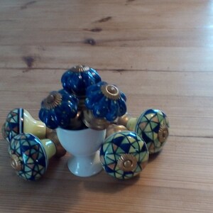 Puede incluir: Un juego de seis pomos de armario de cerámica azul y amarillo con un patrón geométrico. Los pomos están dispuestos en círculo alrededor de una taza de cerámica blanca.