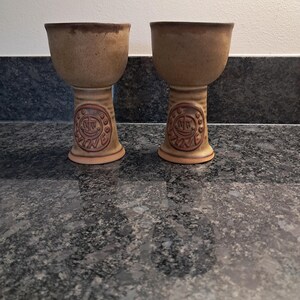 Pode incluir: Dois cálices de cerâmica de cor bronzeada com uma taça larga e base alargada. Cada cálice apresenta um medalhão circular em relevo no caule contendo o texto WINE rodeado por pequenos pontos. A argila tem uma textura salpicada com bordas castanhas.