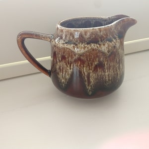 Puede incluir: Una jarra de crema de cerámica marrón y blanca con asa. La jarra tiene un esmalte jaspeado con vetas marrones y blancas.
