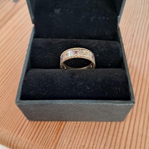 May include: A gold band ring featuring a series of engraved star motifs, each centred with a small, round, vibrant pink gemstone. The ring is presented inside a black velvet-lined jewellery box, showcasing its detailed metalwork and stones.