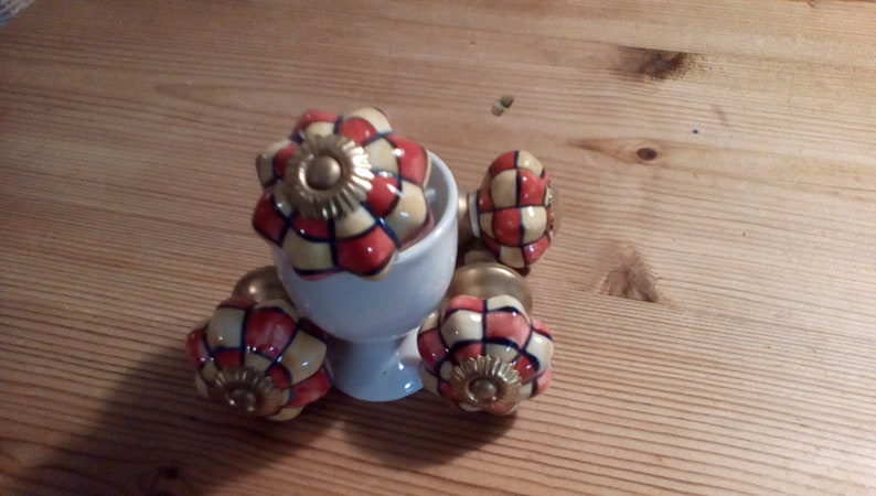 May include: Four ceramic cabinet knobs with a yellow, red, and blue checkered pattern. The knobs have a gold-colored metal base and are displayed on a white ceramic stand.