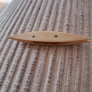 Könnte beinhalten: Ein antiker, cremefarbener Webeschiffchen aus Holz in Bootsform mit zwei kleinen, dunklen Stiften oben. Es liegt auf einem strukturierten, hellfarbigen Stoff, wahrscheinlich Leinen oder Baumwolle.