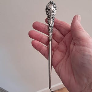 May include: A silver-coloured metal button hook featuring an ornate, embossed handle with intricate scrollwork and floral patterns. The long, slender shaft transitions into a curved hook at the bottom, designed for fastening buttons on clothing items.