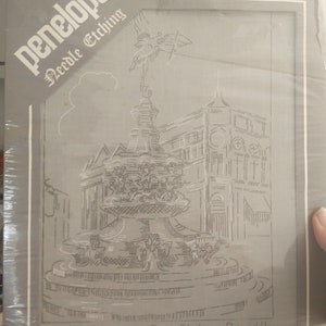 May include: A black and white needlepoint design of Eros Statue - Piccadilly. The design is framed in a gray frame with the word "penelope" and "Needle Etching" in white lettering.