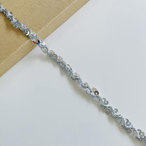 Puede incluir: Un ribete de lentejuelas plateadas con un diseño ondulado. El ribete está hecho de pequeñas lentejuelas redondas y brillantes cosidas a un hilo plateado. El fondo es blanco y beige.