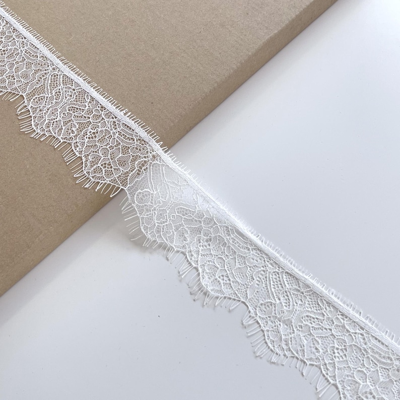 May include: Close-up of a white lace trim with a delicate floral pattern and scalloped edges. The lace is positioned diagonally against a neutral background, showcasing its intricate design and texture. Ideal for sewing and craft projects.