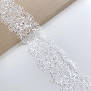 May include: Close-up of a white lace ribbon with a floral pattern. The ribbon has a scalloped edge and is displayed on a white surface with a brown cardboard background. This decorative lace is suitable for various craft projects.