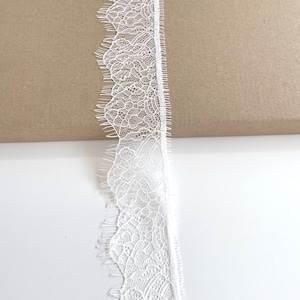 May include: Close-up of a white lace trim with a scalloped edge. The lace has a delicate, intricate floral pattern. The trim is against a white and brown background.