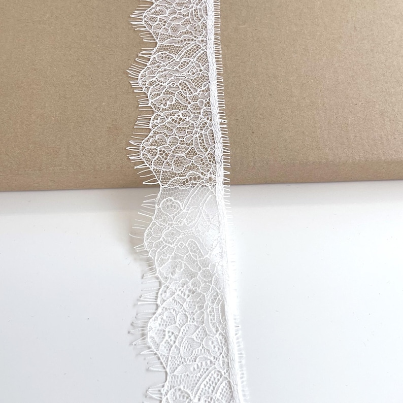 May include: Close-up of a white lace trim with a scalloped edge. The lace has a delicate, intricate floral pattern. The trim is against a white and brown background.