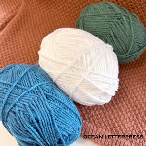 May include: Three balls of yarn in blue, white, and green are arranged on a textured, peach-colored surface. The yarn is tightly wound, and the image includes the text "OCEAN LETTERPRESS".