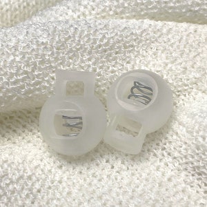 May include: Two translucent white cord locks with a metal spring inside. The cord locks are round with a rectangular opening at the top and a slot for the cord to pass through. The background is a white, textured fabric.
