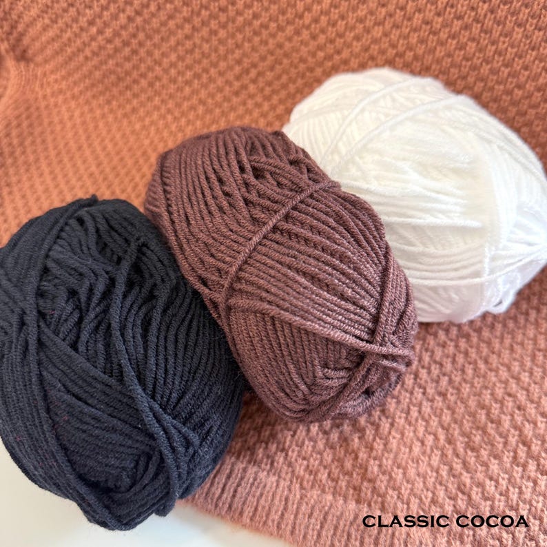 May include: Three balls of yarn in navy blue, cocoa brown, and white are arranged on a textured, cocoa-colored surface. The yarn is tightly wound, and the image includes the text "CLASSIC COCOA".