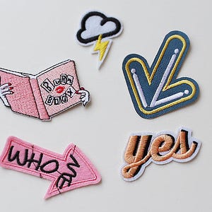 Könnte beinhalten: Fünf gestickte Bügelbilder mit verschiedenen Designs. Die Patches beinhalten ein rosa Buch mit dem Text "Blurb Book" und einer roten Lippe, einen rosa Pfeil mit dem Text "who are", einen blauen Pfeil, ein pfirsichfarbenes Wort "yes" und eine weiße Wolke mit einem gelben Blitz.
