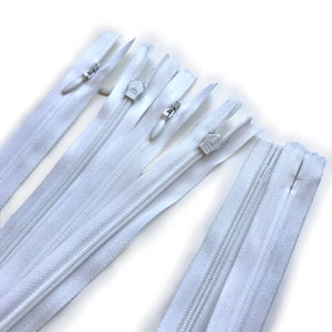 May include: Several white zippers are arranged diagonally against a white background. The zippers have silver metal pulls and teeth. The fabric of the zippers is a bright white color. The zippers are of varying lengths and are new.