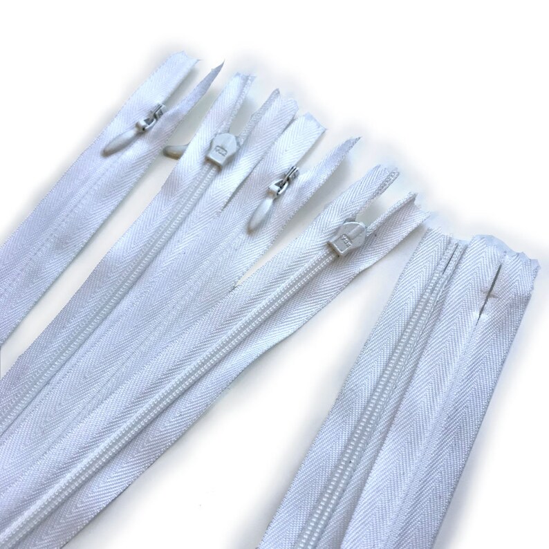 May include: Several white zippers are arranged diagonally against a white background. The zippers have silver metal pulls and teeth. The fabric of the zippers is a bright white color. The zippers are of varying lengths and are new.