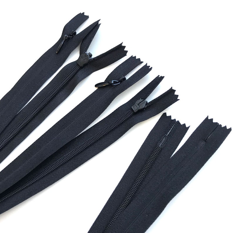 May include: Several black zippers are arranged diagonally against a white background. The zippers are closed, with the zipper pulls visible. The fabric of the zippers appears to be a sturdy, woven material. The image is well-lit, highlighting the details of the zippers.