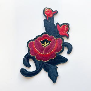 May include: Embroidered floral patch featuring a large, deep red peony with gold accents and a black background. The patch includes two red buds and black leaves, creating a striking contrast against the white background.