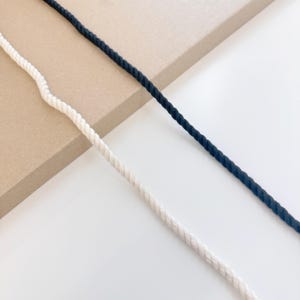 Puede incluir: Primer plano de dos cuerdas, una de color crema y otra azul marino. Las cuerdas están trenzadas y tienen un aspecto texturizado. El fondo es una superficie de color beige claro y blanco.