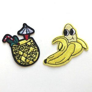 Könnte beinhalten: Zwei gestickte Bügelbilder, eines mit einer Ananas mit einem Cocktail-Schirm und das andere mit einer lächelnden Banane.