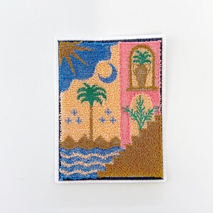 May include: A rectangular, embroidered patch featuring a colorful landscape scene. The design includes a blue sky with a sun and moon, a palm tree, and a pink building with a window. The patch is bordered with white stitching.