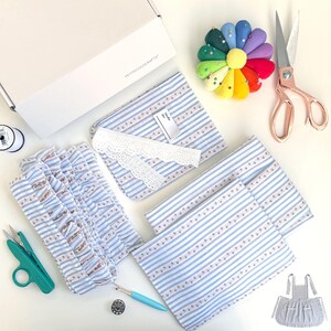 Puede incluir: Un kit de costura para vestido de muñeca con tela a rayas azul y blanca, encaje blanco y ribete de volantes. El kit también incluye un alfiletero, tijeras y una pequeña bobina de hilo. El kit está empaquetado en una caja blanca con el texto "PETITECOOCRAFTS" impreso en ella.