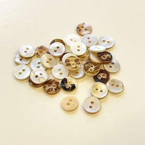 May include: A collection of white and brown shell buttons with two holes each. The buttons are arranged in a pile on a white surface.
