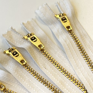 May include: Four white zipper tapes with gold metal zippers. The zippers have a pull tab with the word "K.F.R." printed on them.