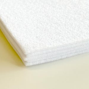 Op de afbeelding: Een stapel witte vellen vilt. Het vilt heeft een zacht, getextureerd oppervlak en is netjes opgestapeld, waarbij de randen van elke laag zichtbaar zijn. De kleur is helder, schoon wit, wat duidt op een ambachtelijke of stoffenvoorraad.