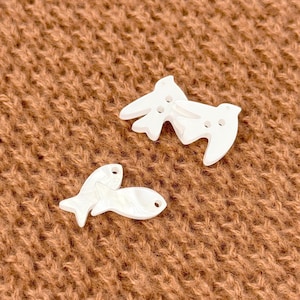 May include: Three white, pearlescent buttons in the shape of a bird and a fish. The bird buttons have two holes, and the fish button has one hole. The buttons are on a brown, textured surface.