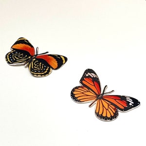 Könnte beinhalten: Zwei bunte Schmetterlingsaufkleber mit orangefarbenen, schwarzen und gelben Akzenten. Die Schmetterlinge befinden sich auf einem weißen Hintergrund.