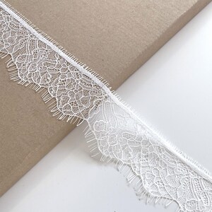 May include: Close-up of white lace trim with a delicate floral pattern and scalloped edge. The lace is on a brown surface. This trim is suitable for various sewing and craft projects.