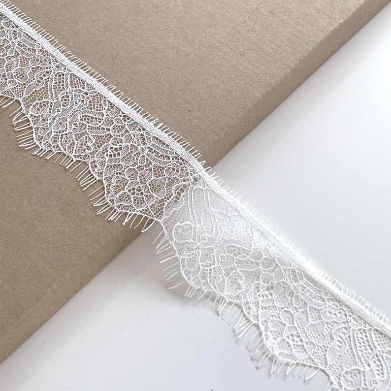 May include: Close-up of white lace trim with a delicate floral pattern and scalloped edge. The lace is on a brown surface. This trim is suitable for various sewing and craft projects.