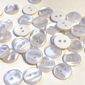 May include: A collection of white iridescent buttons with two holes each. The buttons are scattered on a white surface.