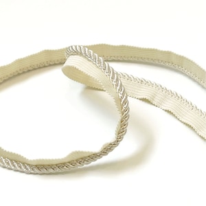 May include: A close-up of a cream-colored, braided trim with a smooth, flat backing. The trim is folded in half, showing both sides.