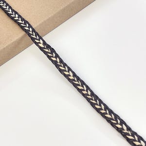 Puede incluir: Primer plano de un cordón de cuero trenzado marrón oscuro con un patrón de chevrón crema. El cordón está colocado en diagonal sobre una superficie blanca y un borde de cartón marrón.