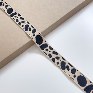 Op de afbeelding: Een beige lint met een zwart dierenprintpatroon. Het lint ligt op een bruin oppervlak.