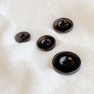 May include: Four black buttons of different sizes on a white knitted fabric. The buttons have four holes each.